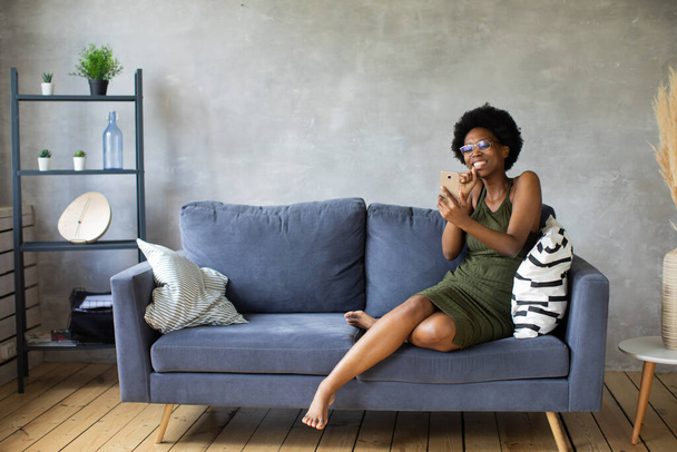 Happy african American young woman sit relax on cozy couch happy to move to new apartment. Smiling black girl rest on comfortable sofa in living room dreaming - 写真・画像