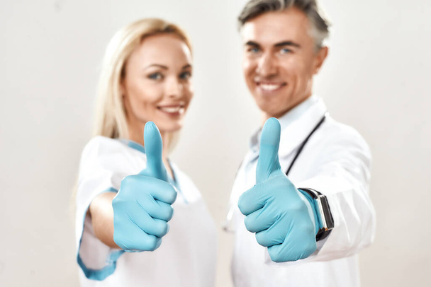 Two happy male and female doctors in medical uniform and blue gloves showing thumbs up, looking at camera and smiling, standing against grey background - 写真・画像