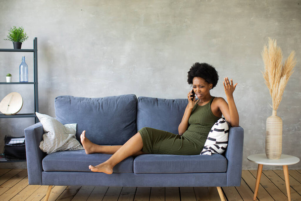 Mujer afroamericana infeliz hablando por teléfono, chica enojada hablando por teléfono, dispositivo móvil descargado o roto, problema con el teléfono, sentado en el sofá - Foto, Imagen