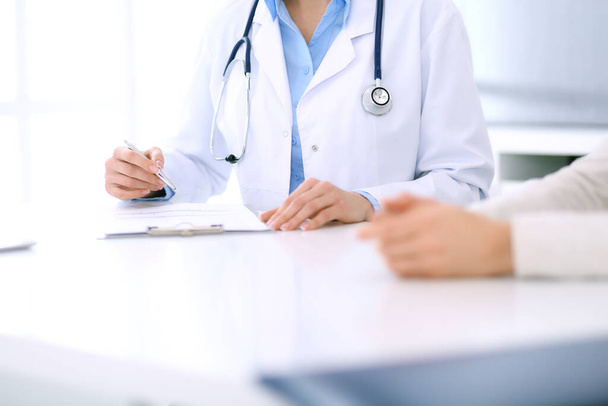 Woman doctor and patient sitting and talking at medical examination at hospital office, close-up. Physician filling up medication history records. Medicine and healthcare concept - Foto, imagen