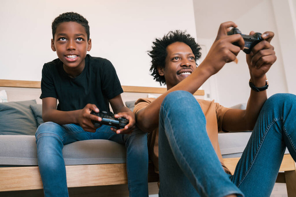 幸せなアフリカ系アメリカ人の父親と息子がソファに座って、家庭でコンソールビデオゲームを一緒にプレイする姿。家族と技術の概念. - 写真・画像