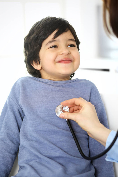 Doctor examining a child patient by stethoscope. Cute arab boy at physician appointment. Medicine and healthcare concept - 写真・画像