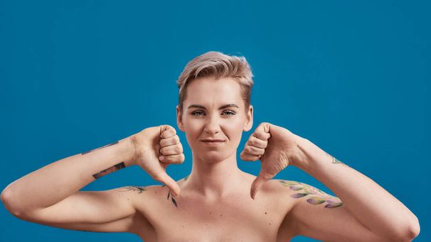 Portrait of a young attractive half naked tattooed woman with perfect skin looking dissatisfied, showing thumbs down isolated over blue background - Photo, image
