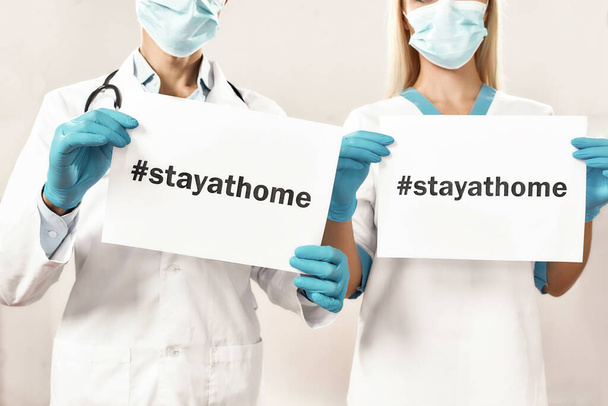 Cropped shot of two doctors wearing protective masks and gloves showing at camera paper sheet with text STAY AT HOME, standing against grey background - Foto, Bild