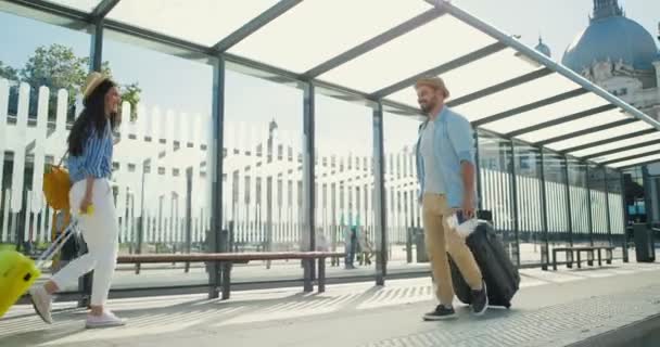 Běloch šťastný pohledný stylový muž kráčející venku na autobusové zastávce a nesoucí kufr na kolečkách. Veselý mužský turista míjí krásnou cestovatelku. Turisté se na sebe usmívají. - Záběry, video