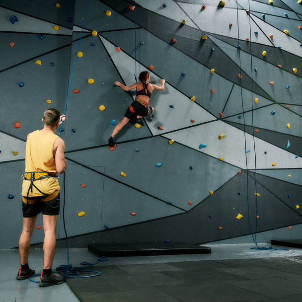Istruttore maschio in possesso di corda, insegnamento, guardando la donna in attrezzature di sicurezza e imbracatura mentre si allena sulla parete di arrampicata artificiale - Foto, immagini