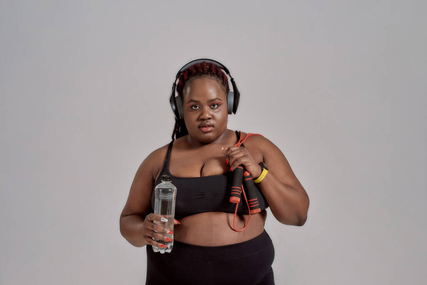 A fresh start. Plump, plus size african american woman in sportswear and headphones holding jump rope and bottle of water, posing in studio over grey background - Photo, image