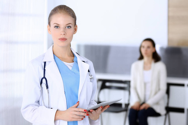 Doctor woman filling up medication history record while standing in emergency hospital office with patient in queue at background. Physician at work, portrait shoot. Medicine and health care concept - Foto, Imagen