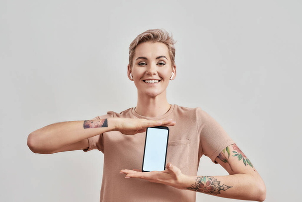 Portrait of tattooed woman with pierced nose and short hair in wireless earphones smiling at camera, advertising, holding smartphone with blank screen isolated over light background - Foto, afbeelding