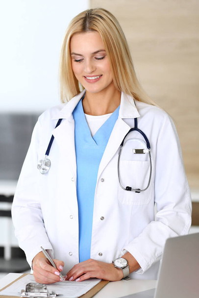 Doctor woman at work in hospital excited and happy of her profession. Blonde physician controls medication history records and exam results. Medicine and healthcare concept - Photo, Image