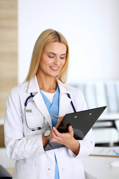 Doctor woman at work in hospital excited and happy of her profession. Blonde physician controls medication history records and exam results while using clipboard. Medicine and healthcare concept - Foto, immagini