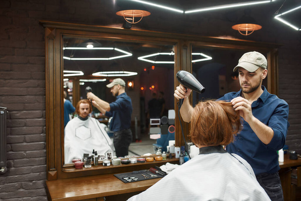 Working in barbershop. Young professional barber drying long redhead hair of his client sitting in armchair in front of the mirror in the modern barbershop - Foto, afbeelding