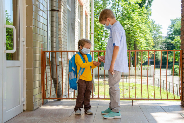 Kinder, Schüler - ein Junge und ein Mädchen stehen vor der Tür der Schule, ein älterer Bruder behandelt die Hände des Mädchens während der Quarantäne des Coronovirus mit einem Desinfektionsspray. - Foto, Bild