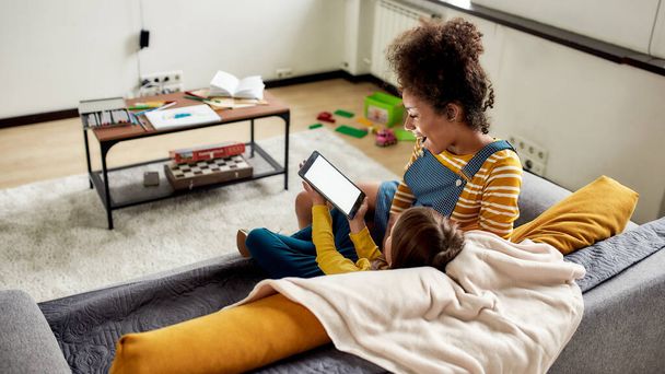 Little Wonder. African american woman baby sitter entertaining caucasian cute little girl. Kid is showing something using tablet pc, sitting on the couch - Photo, Image