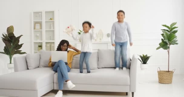 Симпатичные маленькие афроамериканские дети, мальчик и девочка играют и прыгают на диване в гостиной. Мать работает за ноутбуком и сидит на диване рядом. Счастливая мама и шумные дети. - Кадры, видео