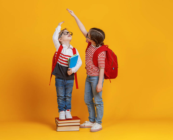 Μικρό αγόρι στέκεται σε στοίβα από βιβλία και προσπαθεί να φτάσει το χέρι του ψηλού κοριτσιού, ενώ ετοιμάζεται να πάει στο σχολείο μαζί στο κίτρινο backdro - Φωτογραφία, εικόνα