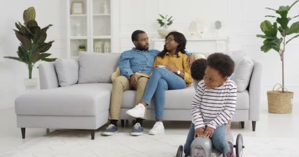 Aranyos kis afro-amerikai gyerekek játszanak a nappaliban. Anya és apa ölelés a kanapén a háttérben. Egy kisfiú, aki csinos lányon lovagol a játékautóban. Otthon. Együtt a család. - Felvétel, videó