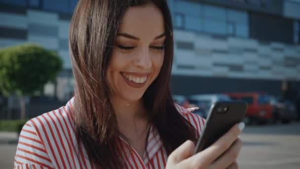 Close up of smiling woman with brunette hair using smartphone outdoors on city background. Beautiful young business female using mobile apps holding smart phone - Footage, Video