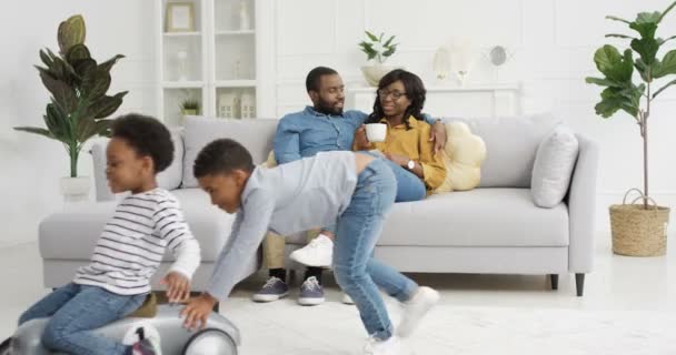 Bonito crianças afro-americanas pequenas brincando na sala de estar. Mãe e pai sentados em abraços com café no sofá no fundo. Menino montando irmã em carro de brinquedo. Em casa. Família em conjunto. - Filmagem, Vídeo