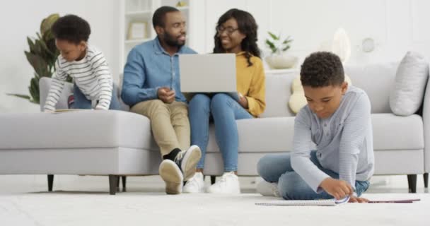 Kis aranyos afro-amerikai gyerekek játszanak és festenek képeket a szülőkkel a hangulatos nappaliban. A szülők kisfiúval és kislánnyal rajzolnak otthon. Együtt töltött családi idő. - Felvétel, videó