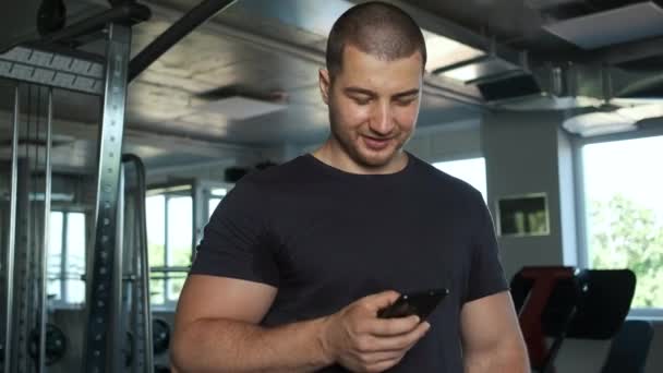 Young healthy brunette athlete standing in a gym and holding a phone in his hands records a workout for a client - Video