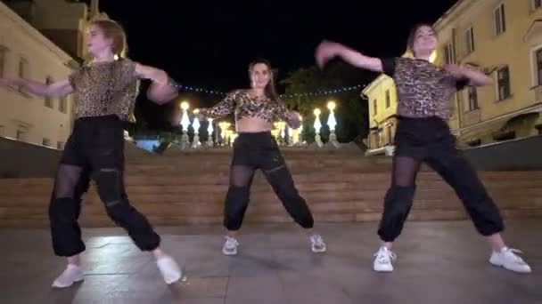 τρία νεαρά χαρούμενα κορίτσια με λεοπαρδαλέ t-shirts χορεύουν στο δρόμο μπροστά από μια παλιά τεράστια πέτρινη σκάλα το βράδυ, εκτελούν μοντέρνους χορούς. Σφηνάκι. - Πλάνα, βίντεο