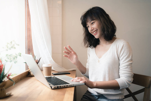 Jovem mulher asiática trabalhando e videoconferência com computador portátil. Uma mulher feliz com um rosto sorridente a trabalhar em casa. Uma chávena de café na mesa de madeira. Trabalho a partir do conceito de casa. Bangkok, Tailândia
 - Foto, Imagem