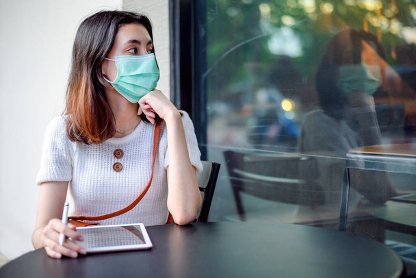 Азиатская бизнесвумен, сидящая в хирургической маске, пользуется таблетками во время пандемии коронавируса-19. - Фото, изображение