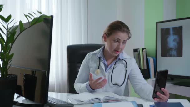 medicina online, medico femminile comunica con il paziente sul cellulare, guarda i risultati dei test sullo schermo del monitor, seduto a un tavolo in ufficio - Filmati, video