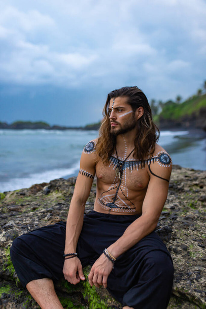 Ein junger, gutaussehender und mutiger europäischer Mann mit Zeichnungen auf seinem Körper sitzt auf dem Hintergrund von Meer und Felsen - Foto, Bild