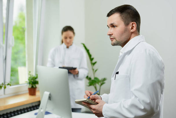 Bělošský lékař si dělá poznámky s perem poblíž ženské terapeutky se schránkou v blízkosti jednoho počítače v nemocnici. - Fotografie, Obrázek