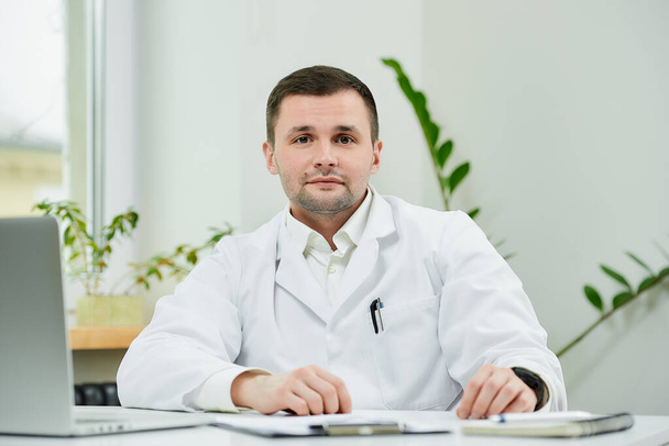 A kind caucasian doctor in a white lab coat is sitting relaxed at his desk and listening in a hospital. A therapist near a laptop is waiting for a patient in a doctor's office. - Photo, Image