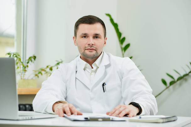 Un buen médico caucásico con una bata blanca de laboratorio está sentado relajado en su escritorio y escuchando atentamente en un hospital. Un terapeuta cerca de un portátil está esperando a un paciente en el consultorio de un médico. - Foto, imagen