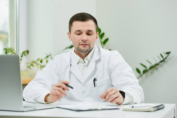 Biały lekarz w białym fartuchu siedzi przy biurku i opuszcza oczy podczas badania historii choroby pacjenta w szpitalu. Terapeuta w pobliżu laptopa w gabinecie lekarskim.. - Zdjęcie, obraz