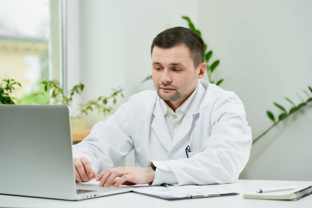 A serious caucasian doctor in a white lab coat is entering the patient's medical history into a computer in a hospital. A therapist is sitting at a desk with a laptop in a doctor's office. - Foto, immagini