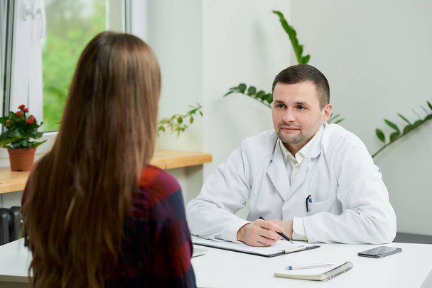 Egy fehér köpenyes fehér orvos ül egy asztalnál, és hallgatja egy női beteg panaszait egy orvosi rendelőben. Egy nő hosszú hajjal egy orvosi vizsgálaton egy kórházban.. - Fotó, kép