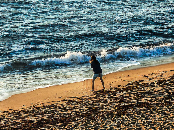 Egy szürke blúzos nő játszik a parton az óceán vizénél naplementekor. - Fotó, kép