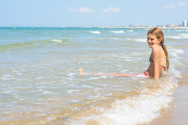 Dziesięciolatka siedzi w wodzie na pustym brzegu morza. - Zdjęcie, obraz
