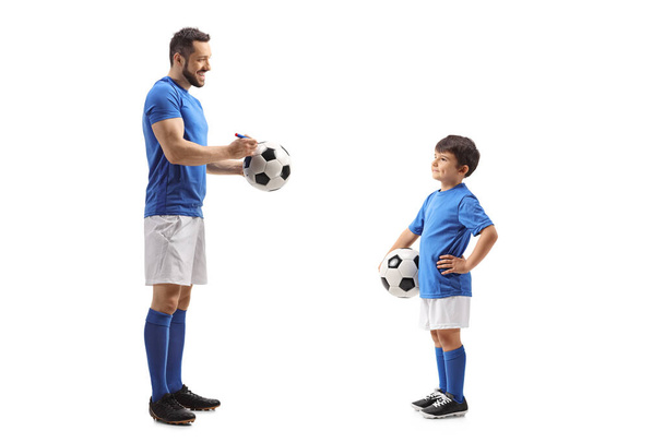 Full length profile shot of a footballer signing an autograph on a soccer ball for a junior football player isolated on white background - Foto, Imagen