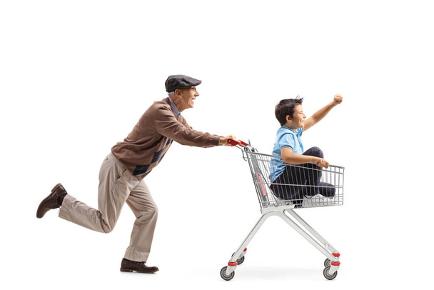 Full length profile shot of an elderly man running and pushing a shopping cart with a boy inside isolated on white background - 写真・画像