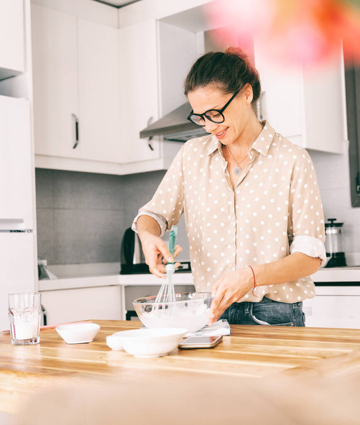 Beautiful young woman in glasses prepares dessert in the kitchen, whips squirrels and dough and smiles happily - Фото, изображение