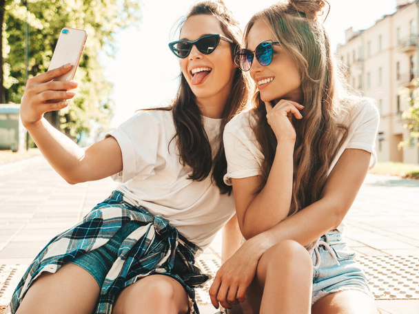 Two young smiling hipster women in summer clothes. Girls taking selfie self portrait photos on smartphone.Models sitting in the street.Female going crazy and making funny faces in sunglasses - Fotó, kép
