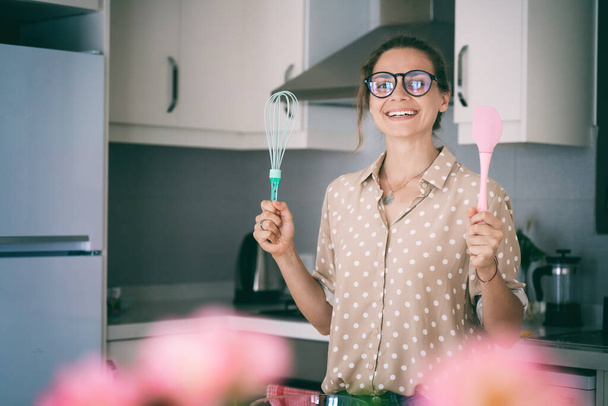 Beautiful young woman in glasses prepares dessert in the kitchen, holds a whisk and a spatula in her hands, and smiles happily. - Φωτογραφία, εικόνα
