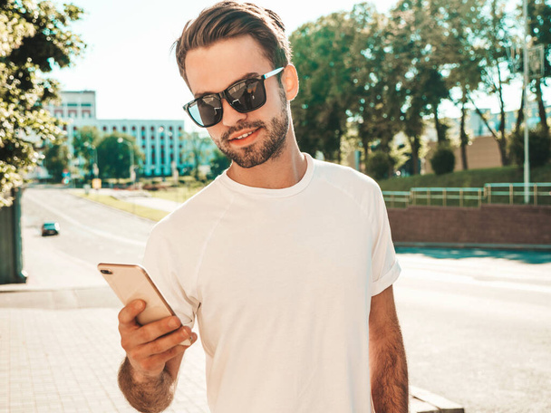 Handsome smiling stylish hipster lambersexual model.Man dressed in white T-shirt.Fashion male posing in the street. Holding, scrolling and texting in his cellphone. Writes sms on smartphone - Photo, Image