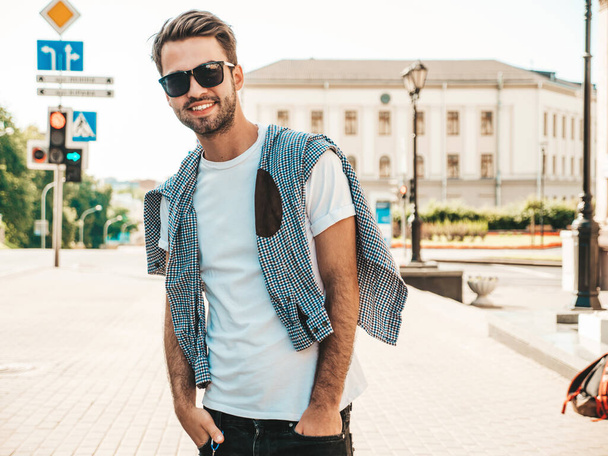 Portrait of handsome smiling stylish hipster lambersexual model.Man dressed in white T-shirt. Fashion male posing on the street background in sunglasses - Foto, imagen