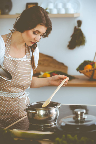 Young brunette woman cooking soup in kitchen. Housewife holding wooden spoon in her hand. Food and health concept - Zdjęcie, obraz