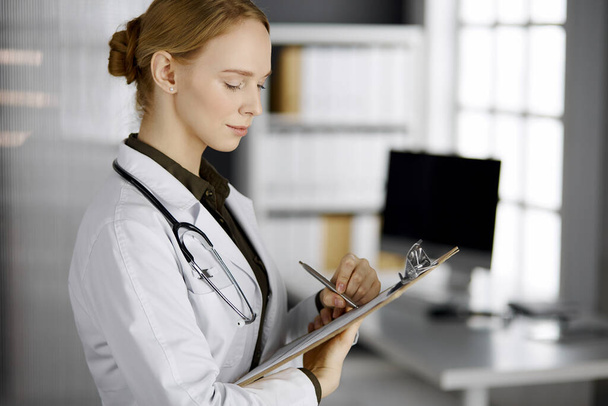 Friendly smiling female doctor using clipboard in clinic. Portrait of friendly physician woman at work. Medical service in hospital. Medicine concept - Photo, Image
