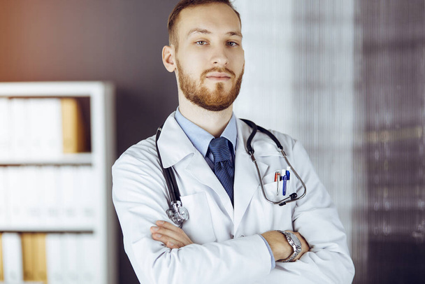 Red-bearded doctor standing straight in sunny clinic near his working place. Portrait of physician. Medicine concept - Foto, afbeelding