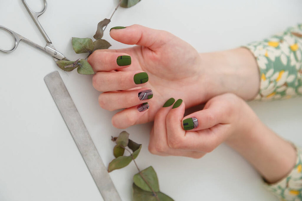 Detailní záběr rukou s geometrickým zeleným designem nehtů. Nůžky, pilníky na nehty a listy eukalyptu na bílém stole. Lak na nehty s gelovým leštěním, na ruce a nehty. - Fotografie, Obrázek