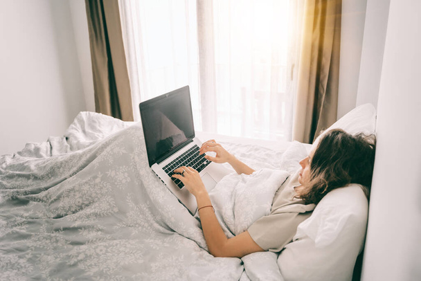 Serious young woman is lying on a bed under a blanket with a laptop in front of her. Problems Remote work. Freelancer concept. - 写真・画像